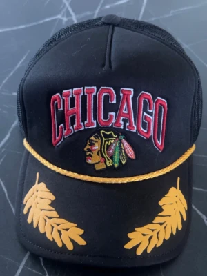 Svart Chicago keps med brodyr - Snygg svart keps med broderad CHICAGO-text i rött och vitt, samt ett färgglatt emblem framtill. Kepsen har gulddetaljer i form av ett snöre och blad på skärmen. Klassisk trucker-modell med mesh baktill.