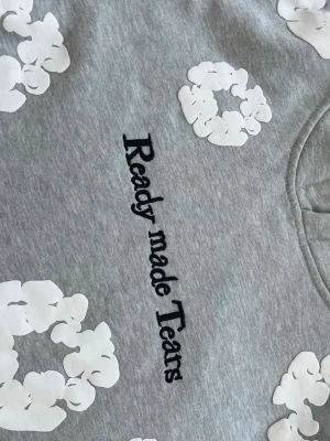 Grå hoodie Ready made Tears - Säljer en grå hoodie från Ready made Tears med vita blommönster tryckta över hela tröjan och svart text på bröstet. Hoodien har en klassisk huva och är gjord i mjukt bomullsmaterial. Perfekt för dig som gillar streetwear och unika prints.