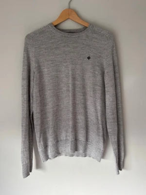 Grå stickad tröja från Morris - Snygg grå stickad tröja från Morris i 100% merinoull. Tröjan har rund halsringning, diskret broderad logga på bröstet och ribbade muddar vid ärmslut och nederkant. Perfekt basplagg för en clean och stilren look.