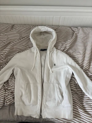Vit hoodie från Polo Ralph Lauren - Snygg vit hoodie från Polo Ralph Lauren med dragkedja, två stora fickor fram och klassisk huva med dragsko. Tillverkad i mjuk bomullsblandning och har den ikoniska lilla loggan broderad på bröstet. Perfekt för en chill och stilren look.