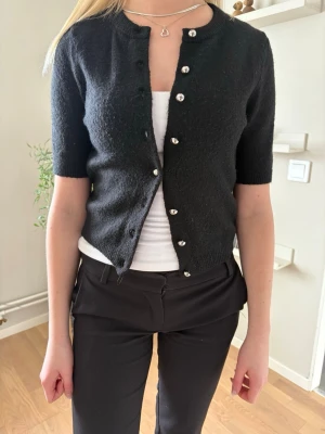 Svart kofta med kort ärm och pärlknappar - Stilren svart kofta med korta ärmar och dekorativa pärlknappar framtill. Koftan har rund halsringning och är gjord i ett mjukt, stickat material. Perfekt att bära över ett linne eller t-shirt för en enkel men snygg look.