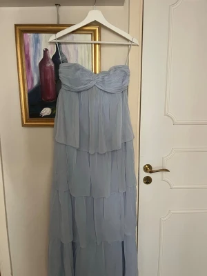 Ljusblå balklänning med volanger - Romantisk ljusblå långklänning med flera lager volanger. Klänningen har en söt byst med twistad detalj framtill och är tillverkad i ett lätt, luftigt material som ger ett drömmigt intryck. Perfekt för bal och för fina teman. Använt en gång och inga täcken på använding