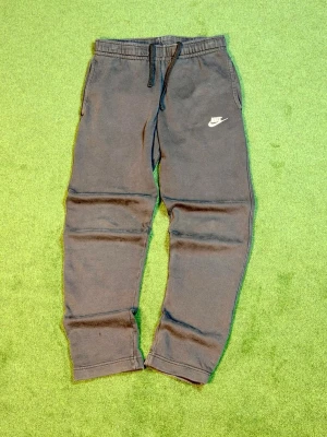 Svarta mjukisbyxor från Nike - Säljer ett par svarta mjukisbyxor från Nike med dragsko i midjan och resår vid bensluten. Byxorna har två sidofickor och en vit Nike-logga på vänster ben. Perfekta för chill dagar eller träning.