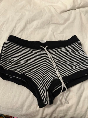 Svartvita randiga shorts med snörning - Säljer ett par svartvita shorts med tunna horisontella ränder och vit dragsko i midjan. Shortsen har bredare svarta kanter i midja och benslut, samt en mjuk och stretchig passform. Perfekta för en avslappnad och sportig stil.