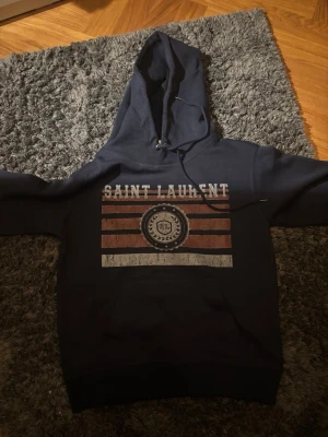 Mörkblå hoodie från Saint Laurent - Snygg mörkblå hoodie från Saint Laurent med stor huva och dragsko. Framsidan har ett tryck med texten 'SAINT LAURENT' och grafiska ränder i vitt och rött. Tillverkad i mjukt bomullsmaterial som känns skönt mot huden. Perfekt för en avslappnad och trendig look.