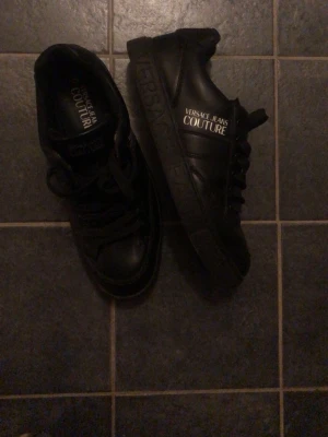 Svarta Versace Jeans Couture sneakers - Snygga svarta sneakers från Versace Jeans Couture med logga i vitt på sidan och plösen. Skorna har klassisk låg profil, svarta skosnören och är tillverkade i slätt konstläder. Perfekta för dig som vill ha en stilren och trendig look.