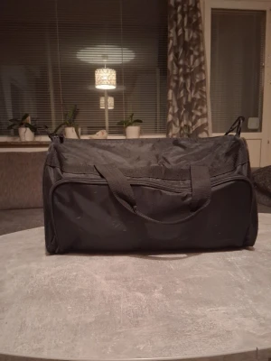 Svart sportbag med dragkedja - En enkel och rymlig svart sportbag i tygmaterial med dubbla handtag och dragkedja. Väskan har en avlång form och stor öppning för enkel packning. Perfekt för gymmet eller som weekendbag. Inga synliga loggor eller märken.