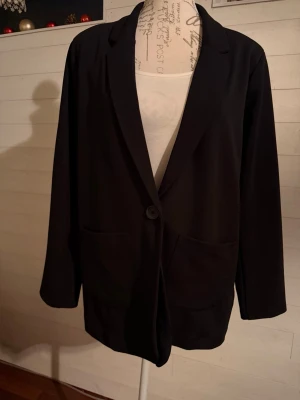 Svart enkel kavaj med knapp - Stilren svart kavaj med en knapp framtill och två stora fickor. Klassisk passform med smala slag och lång ärm. Perfekt för en clean och modern look. Materialet ser ut att vara mjukt och lätt, troligen polyester eller bomullsblandning.