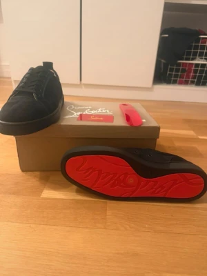 Svarta Christian Louboutin - Säljer ett par svarta låga sneakers från Christian Louboutin med klassisk röd sula och diskret logga undertill. Skorna har svart textil och mockaliknande material, svarta skosnören och metallöljetter på sidan. Perfekta för dig som gillar stilrena och exklusiva sneakers.