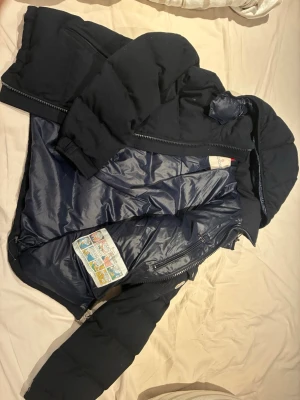  Jacka Moncler - Snygg mörkblå dunjacka från Moncler med avtagbar huva och dragkedja framtill. Jackan har en ficka med Moncler-logga på ärmen och två sidofickor med dragkedja. Insidan är glansig och quiltad för extra värme. Perfekt för kalla vinterdagar. Passar L/M. 