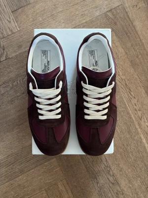 Vinröda Maison Margielas  - Snygga sneakers från Maison Margiela i bordeauxrött och mörkbrunt läder och mocka. Klassisk low-top modell med vita snören och gummisula. Perfekta för dig som gillar stilrena och trendiga skor med en lyxig touch.