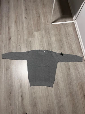 Grå stickad tröja från Stone Island - Snygg grå stickad tröja från Stone Island med rund halsringning och ribbade muddar. Tröjan har en enkel och stilren design som passar perfekt till jeans eller chinos. Tillverkad i mjukt material för extra komfort.