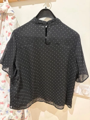 Svart blus med små vita prickar - Snygg svart blus med små vita prickar och korta ärmar. Blusen har en hög krage med knappstängning i nacken och är gjord i ett tunt, lätt och något transparent material. Perfekt för en trendig och avslappnad look.