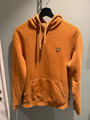 Orange hoodie från Lyle & Scott - Snygg orange hoodie från Lyle & Scott med klassisk gul logga på bröstet. Hoodien har huva med dragsko och en stor magficka framtill. Tillverkad i mjukt bomullsmaterial och har ribbade muddar vid ärmar och nederkant. Perfekt för en avslappnad och trendig look. Storlek small.
