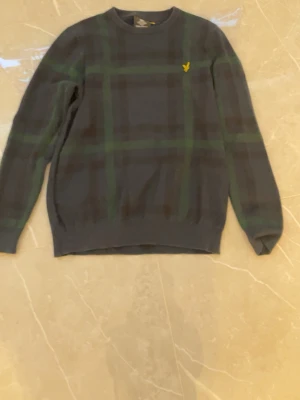 Mörkblå rutig stickad tröja Lyle & Scott - Stickad tröja från Lyle & Scott i mörkblått med grönt och brunt rutmönster. Klassisk rund halsringning och gul logga broderad på bröstet. Perfekt för dig som gillar stilrena och tidlösa plagg med en twist.