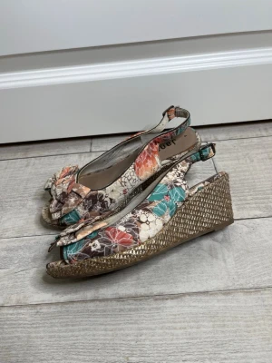 Blommiga kilklackssandaler från Jana - Säljer ett par färgglada sandaler från Jana med blommigt mönster i orange, turkos, beige och brunt. Sandalerna har öppen tå, rosett framtill och justerbar rem i hälen. Kilklack i flätad look ger extra höjd och sommarkänsla. Perfekta för dig som vill sticka ut.