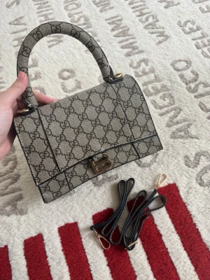 Gucci beige monogram handväska - Snygg beige handväska från Gucci med klassiskt GG-monogram över hela väskan. Väskan har ett topphandtag, guldfärgade detaljer och medföljande axelremmar i matchande material. Tillverkad i canvas med läderdetaljer och elegant metallspänne framtill.