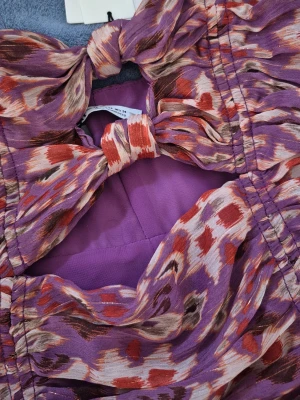 Lila mönstrad kort klänning med rosett - Supersnygg kort klänning i lila med färgstarkt mönster i rött, vitt och svart. Klänningen har en stor rosettdetalj framtill, rynkade sidor och en cut-out vid bysten. Perfekt för dig som vill sticka ut! Materialet känns lätt och luftigt. Zara, helt ny