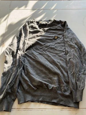 Grå stickad tröja från Lyle & Scott - Snygg grå stickad tröja från Lyle & Scott med klassisk gul logga på bröstet. Tröjan har v-ringning, långa ärmar och ribbade muddar vid ärmslut.