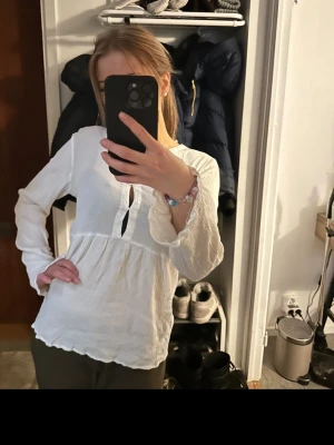 Vit blus med volang - Säljer en söt vit blus med v-ringning och dekorativ knappdetalj framtill. Blusen har långärmade ärmar med lätt utsvängda muddar och ett luftigt, krinklat tyg som ger en avslappnad känsla. Perfekt till jeans eller kjol för en trendig look.