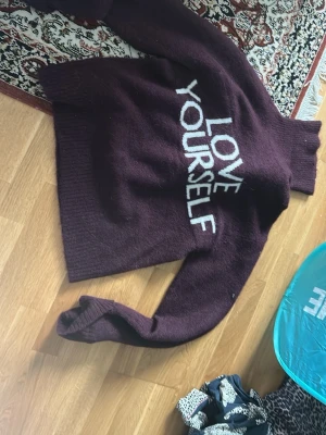 Vinröd stickad tröja Gina Tricot - Mysig vinröd stickad tröja från Gina Tricot med hög krage och texten 'LOVE YOURSELF' i vitt framtill. Tröjan har lång ärm och ribbade muddar vid hals, ärmslut och nederkant. Perfekt för kalla dagar och en snygg statement-piece i garderoben.