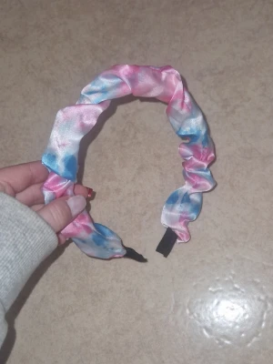 Scrunchie-diadem - Säljer ett scrunchie-diadem i glansigt tyg med pastellfärger i rosa, blått och vitt. Diademet har en rynkad design som ger en trendig och lekfull look. Perfekt accessoar för att piffa upp frisyren med en färgklick.