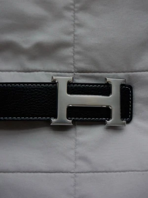 Svart skärp med H-spänne  - Snyggt svart skärp med klassiskt H-format spänne i silverfärg. Skärpet är gjort i präglat skinn och har vita kontrastsömmar längs kanterna. Perfekt accessoar för att lyfta din outfit.