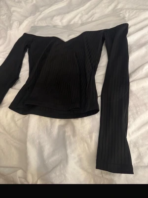 Svart offshoulder topp med ribbad struktur - Trendig svart offshoulder topp med lång ärm och ribbad struktur. Toppen har en snygg v-ringad skärning och en kontrasterande vit kant upptill som ger en cool twist. Perfekt för dig som vill ha en stilren men ändå unik look.