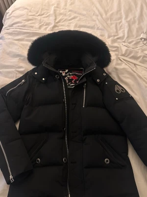 Svart dunjacka från Moose Knuckles äkta - Säljer en svart dunjacka från Moose Knuckles med stor pälskantad huva och silverfärgade detaljer. Jackan har flera dragkedjor, tryckknappar och den ikoniska logotypen på ärmen. Perfekt för kalla vinterdagar och riktigt snygg streetstil.