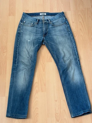 Acne Studios blåa jeans - Säljer ett par klassiska blå jeans från Acne Studios med rak passform och snyggt slitna detaljer. Jeansen har fem fickor, knappgylf och är tillverkade i kraftig denim. Perfekta för en avslappnad och trendig look.