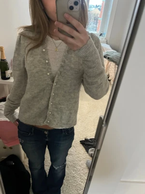 Grå kofta med pärlknappar - Mysig grå kofta med pärlknappar och diskret scallop-detalj vid kragen. Koftan har lång ärm och är stickad i ett mjukt material som passar perfekt till jeans. Enkel och stilren design som är lätt att matcha.(en av knapparna sitter lite löst, men inget som syns)