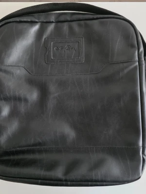 Svart axelremsväska i konstskinn från Björn Borg  - Snygg svart axelremsväska från Björn Borg. Väskan har en stilren design med diskret logotyp framtill och praktiska fack för mobil och tillbehör. Perfekt för dig som vill ha en klassisk och tidlös look till plugget eller jobbet.