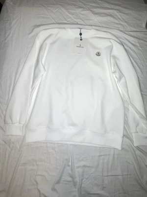 Vit sweatshirt från Moncler - Säljer en stilren vit sweatshirt från Moncler med rund halsringning och Moncler-logga på bröstet. Tröjan har ribbade muddar vid ärmslut och nederkant samt en diskret logga på ärmen. Perfekt för dig som gillar minimalistisk design.