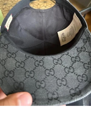 Svart keps från Gucci med GG-mönster - Snygg svart keps från Gucci med det klassiska GG-monogrammet över hela kepsen. Kepsen har böjd skärm och detaljer i rött och grönt på sidorna. Tillverkad i slitstarkt tyg med justerbar passform. Perfekt för dig som vill ha en exklusiv och trendig accessoar.