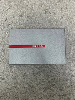 Grå sneakers från Prada - Snygga grå sneakers från Prada med vita snören och detaljer i mesh och glansigt material. Klassisk låg modell med röd logga på plösen och insidan. Perfekta för dig som vill ha en clean och exklusiv look.