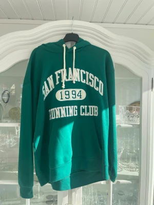 Grön hoodie San Francisco Primark - Grön hoodie från Primark med vit text 'San Francisco Running Club 1994' på bröstet. Klassisk modell med huva och vita dragsnören. Tillverkad i mjukt material, perfekt för en avslappnad och sportig stil.