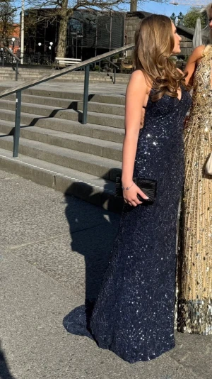 Marinblå glittrig balklänning - Säljer en marinblå långklänning med glittriga detaljer och smala axelband. Klänningen har en figurnära passform, öppen rygg och är täckt av paljetter samt pärlor. Perfekt för bal eller festliga tillfällen. Klädpåse ingår. Endast använd 1 gång.