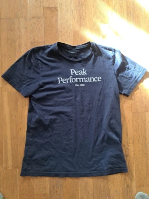 Mörkblå Peak Performance t-shirt - Klassisk  t-shirt från Peak Performance med vit logga och text på bröstet. T-shirten har rund halsringning och korta ärmar. Tillverkad i mjuk bomull som känns skön mot huden. Perfekt för en avslappnad och stilren look.