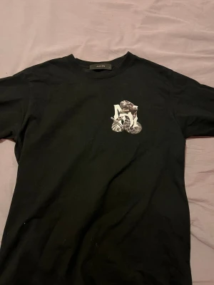 Svart tröja från Amiri med tryck -  STRL S.     Säljer en svart tröja från Amiri med ett unikt tryck på bröstet. Tröjan har rund hals och korta ärmar. Materialet känns mjukt och skönt, troligtvis bomull. Perfekt för dig som gillar streetwear och stilrena plagg. 