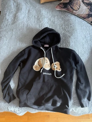 Palm angels hoodie - Säljer en svart hoodie från Palm Angels med ett stort broderat björnmotiv på bröstet och vit logotyptext. Hoodien har huva med vita snören och en stor magficka. Perfekt för dig som gillar streetwear och vill sticka ut.