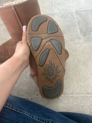 Bruna fårskinnsstövlar UGG - Mysiga bruna stövlar från UGG i äkta fårskinn med mjukt, fluffigt foder. Klassisk modell med rund tå och mellanhögt skaft. Yttermaterialet är mocka och sulan har greppvänliga gummipartier. Perfekta för kalla dagar och enkel att matcha till olika outfits.