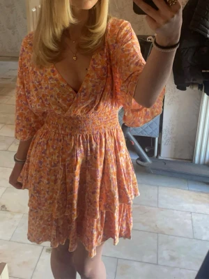 Blommig kortklänning med volang - Söt kortklänning med blommigt mönster i orange, rosa och lila toner. Klänningen har omlott-v-ringning, volangdetaljer och smock i midjan. Ärmarna är trekvarts och tyget känns lätt och luftigt, perfekt för varma dagar.