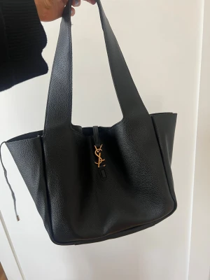 Svart YSL handväska i läder - Stilren svart handväska från Yves Saint Laurent i mjukt, kornigt läder. Väskan har breda axelband och ett elegant YSL-monogram i guld framtill. Rymlig modell med klassisk form och exklusiv känsla, perfekt för dig som gillar lyxiga accessoarer. Osäker på äkthet därav billigt. 