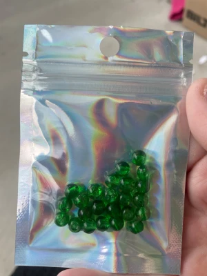 Gröna glaspärlor i påse - Säljer ett gäng klara, runda gröna akrylpärlor i en holografisk zip-påse. Perfekta för att skapa egna smycken som armband, halsband eller örhängen. Pärlorna har en djup grön färg och är genomskinliga vilket ger ett snyggt ljusspel.