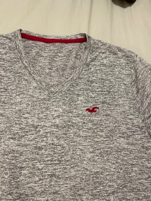 Gråmelerad v-ringad t-shirt Hollister - Säljer en gråmelerad t-shirt från Hollister med v-ringning och korta ärmar. T-shirten har ett diskret rött Hollister-logo broderat på bröstet och är tillverkad i mjuk bomull. Perfekt för en avslappnad och stilren look.