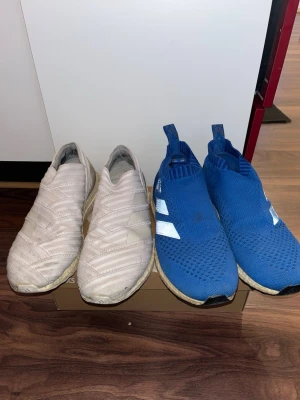 Adidas slip-on sneakers vita (44,2/3)  & blå (43,1/2) - Två par Adidas slip-on sneakers, ett par i vitt med diskreta ränder och ett par i blått med vita detaljer. Båda paren har stickad ovandel och elastisk passform utan snörning. Sulan är vit och ger en sportig vibe. Perfekta för dig som gillar streetwear och enkel stil.