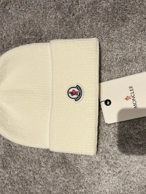 Vit ribbstickad mössa från Moncler(minst 300kr) - Snygg vit ribbstickad mössa från Moncler med uppvikt kant och klassisk Moncler-logga framtill. Tillverkad i mjukt material som håller dig varm och ger en clean look. Perfekt accessoar för kyliga dagar och enkel att matcha med olika outfits.