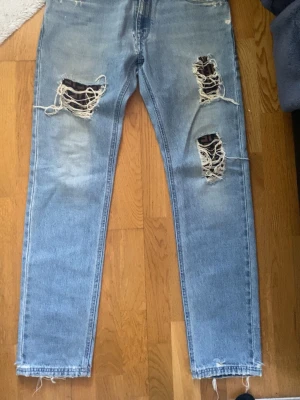 Ljusa ripped jeans från Fendi - Säljer ett par ljusblå jeans från Fendi med kraftiga slitningar och stora hål på båda benen. Jeansen har raka ben och klassisk femficksmodell. Tillverkade i denim med snygg tvätt och råa kanter vid benslut. Perfekta för en trendig och avslappnad stil.