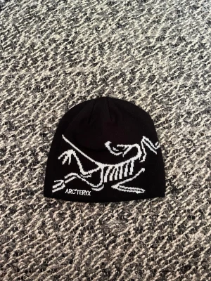 Svart mössa från Arc'teryx - Säljer en svart mössa från Arc'teryx med deras ikoniska vita fossil-logga på framsidan. Mössan är tunn och stretchig, perfekt för kyliga dagar. Materialet känns mjukt och följsamt, och designen är clean med en snygg kontrast mellan svart och vitt.