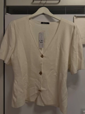 Vit kortärmad tröja från Gina Tricot - Stilren vit tröja från Gina Tricot med v-ringning och tre dekorativa knappar framtill. Kortärmad modell i mjukt material som passar perfekt till både jeans och kjol. Enkel att matcha och ger en fräsch look till din outfit.